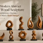 An image titled "10 Modern Abstract Teak Wood Sculpture for Contemporary Homes – Warmth Meets Bold Design." Ten unique, hand-carved abstract teak wood sculptures with rich grain patterns are displayed on black bases across a two-tiered concrete ledge. The pieces showcase organic shapes, including spirals, loops, a crescent moon, and fluid twists, set against a minimalist, warm-toned contemporary living room background.