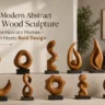 An image titled "10 Modern Abstract Teak Wood Sculpture for Contemporary Homes – Warmth Meets Bold Design." Ten unique, hand-carved abstract teak wood sculptures with rich grain patterns are displayed on black bases across a two-tiered concrete ledge. The pieces showcase organic shapes, including spirals, loops, a crescent moon, and fluid twists, set against a minimalist, warm-toned contemporary living room background.