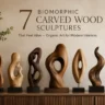 A wide landscape shot of seven biomorphic carved wood sculptures lined up on a fluted wooden console table. Each abstract sculpture showcases unique fluid shapes, cutouts, and distinct natural wood grain tones. On the warm textured wall above, elegant brown typography reads: "7 Biomorphic Carved Wood Sculptures That Feel Alive – Organic Art for Modern Interiors." A ceramic vase with delicate green branches sits on the far left, completing the modern earthy aesthetic.